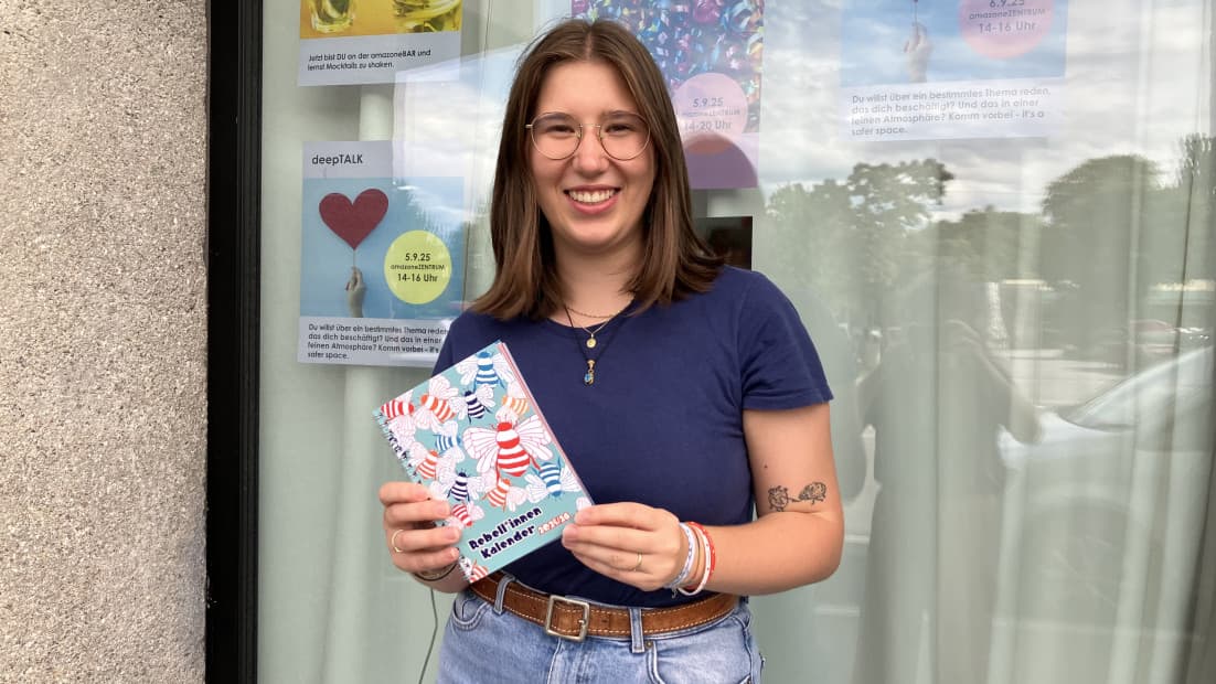 Eine junge Frau hält den Rebell*innen Kalender in den Händen und lächelt in die Kamera.