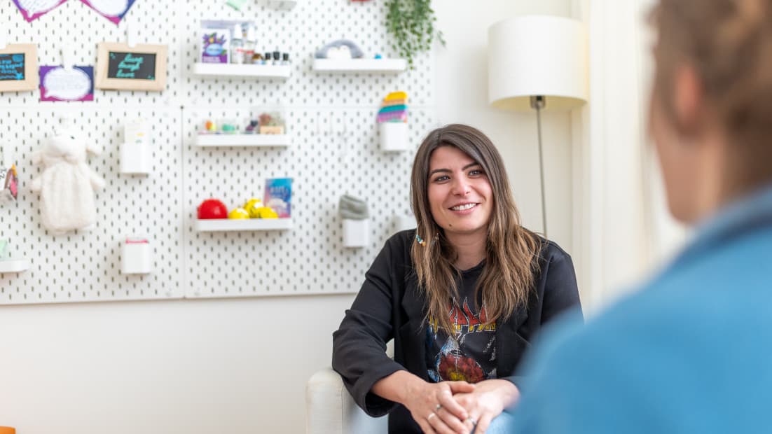 Man sieht über die Schulter einer Person eine weiblich gelesene Person, die dasitzt und lächelt.