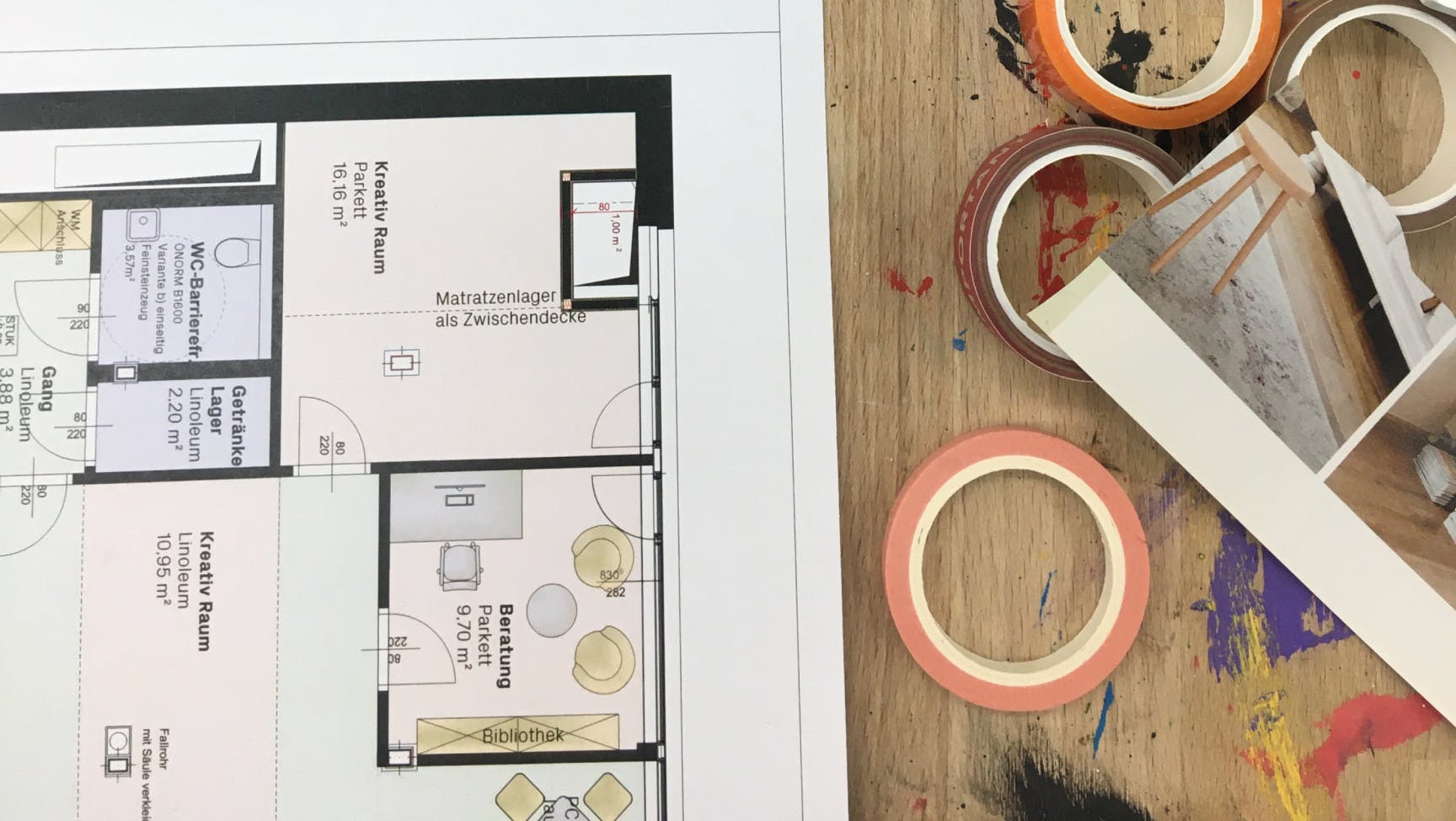 ALT Text: Auf einem Werkstatttisch sieht man einen Teil eines Grundrissplans, unterschiedliche Klebebänder und 2 Behälter mit Wachsmalkreiden liegen.