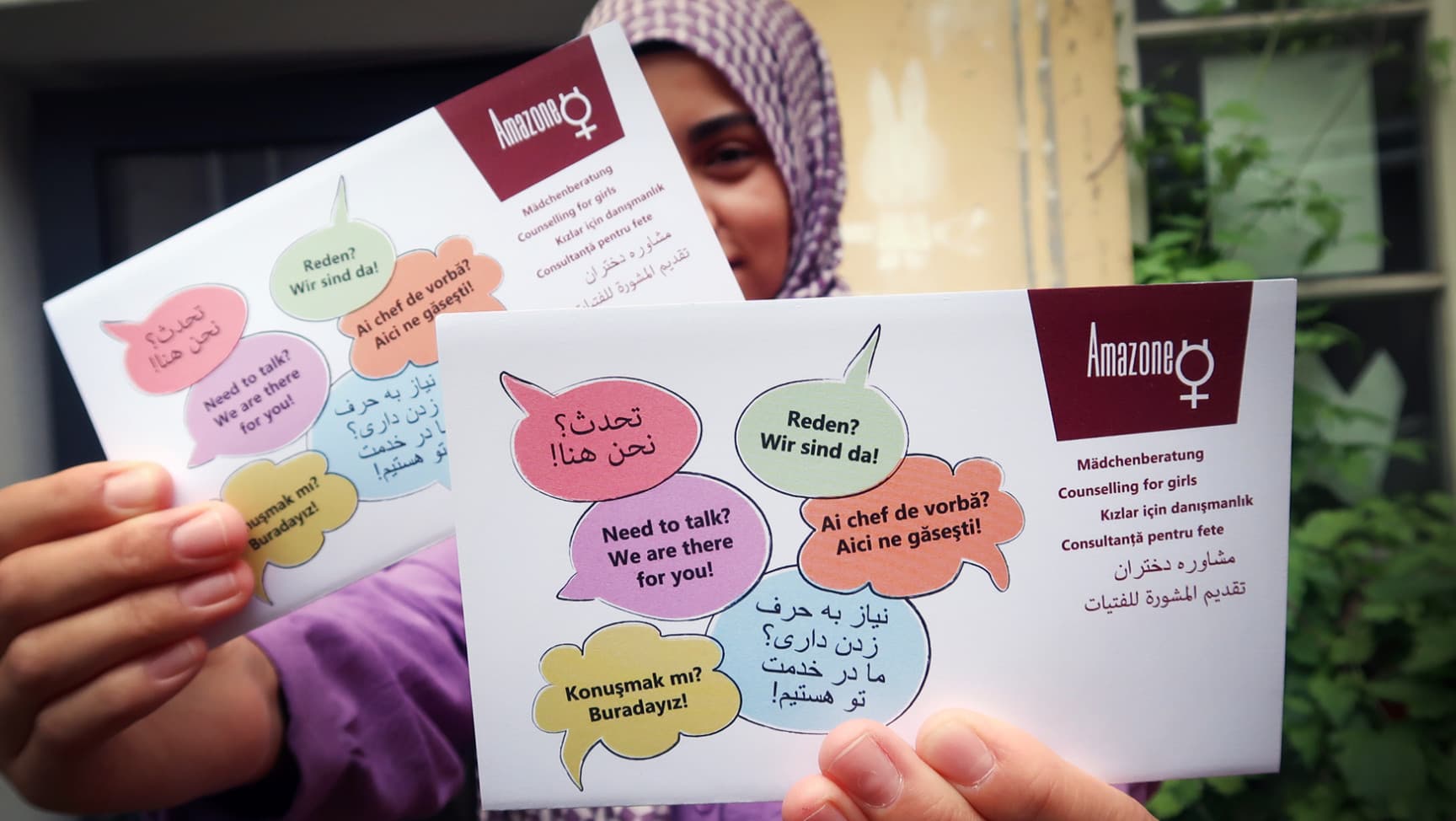 Eine weiblich gelesene Jugendliche hält den mehrsprachigen Flyer des Vereins Amazone in die Kamera.