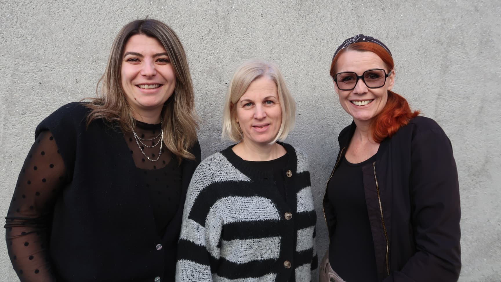 Drei Beraterinnen stehen nebeneinander vor einer grauen Wand und lachen in die Kamera.