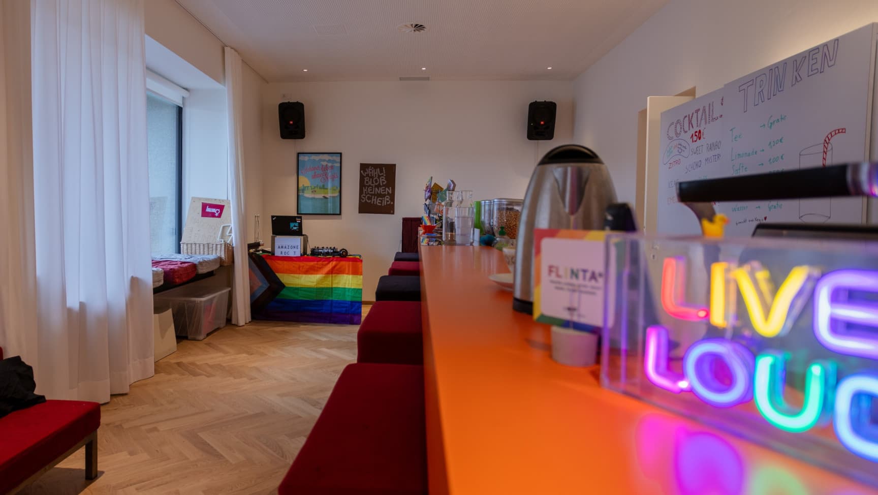 Ein Raum mit einer Bar, Barhockern, ein großes Fenster mit Sitzmöglichkeiten und ein DJane Pult im Hintergrund.