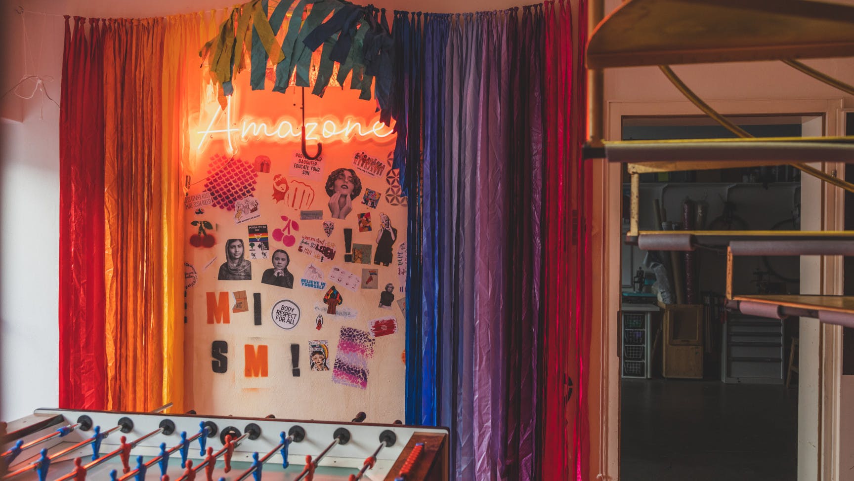 Ein Raum mit einer goldenen Wendeltreppe, ein Tischfußballtisch und eine Wand mit Regenbogenvorhang und Amazone Leuchtschrift