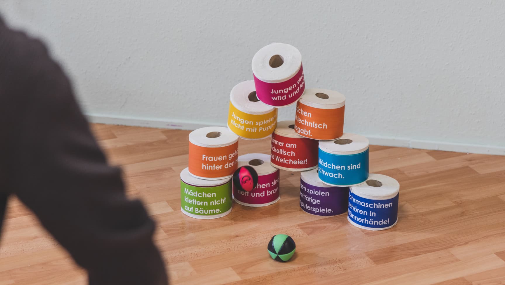 Aufgestapelte Klorollen mit Statements darauf werden mit Bällen abgeschossen.