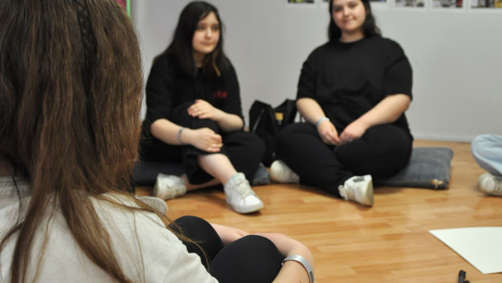 Drei Personen sitzen auf dem Boden. Hinter ihnen sind Bilder an der Wand