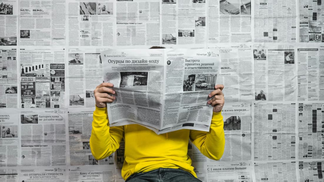 Person sitzt da mit aufgeschlagender Zeitung vor dem Gesicht