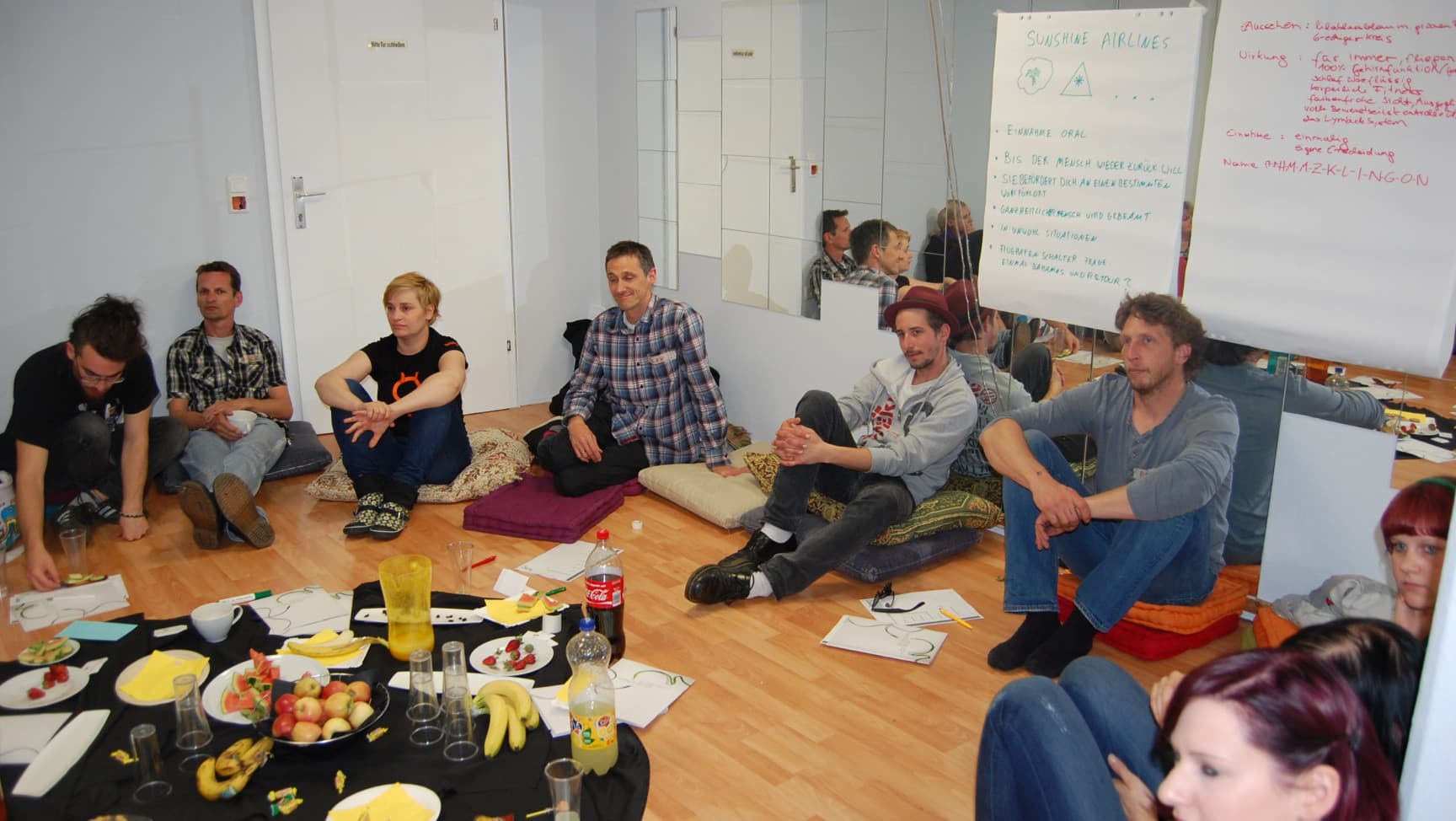 Erwachsene sitzen auf dem Boden im Kreis. Hinter ihnen Flipcharts an der Wand, in der Mitte Essen, Getränke, Arbeitsutensilien.