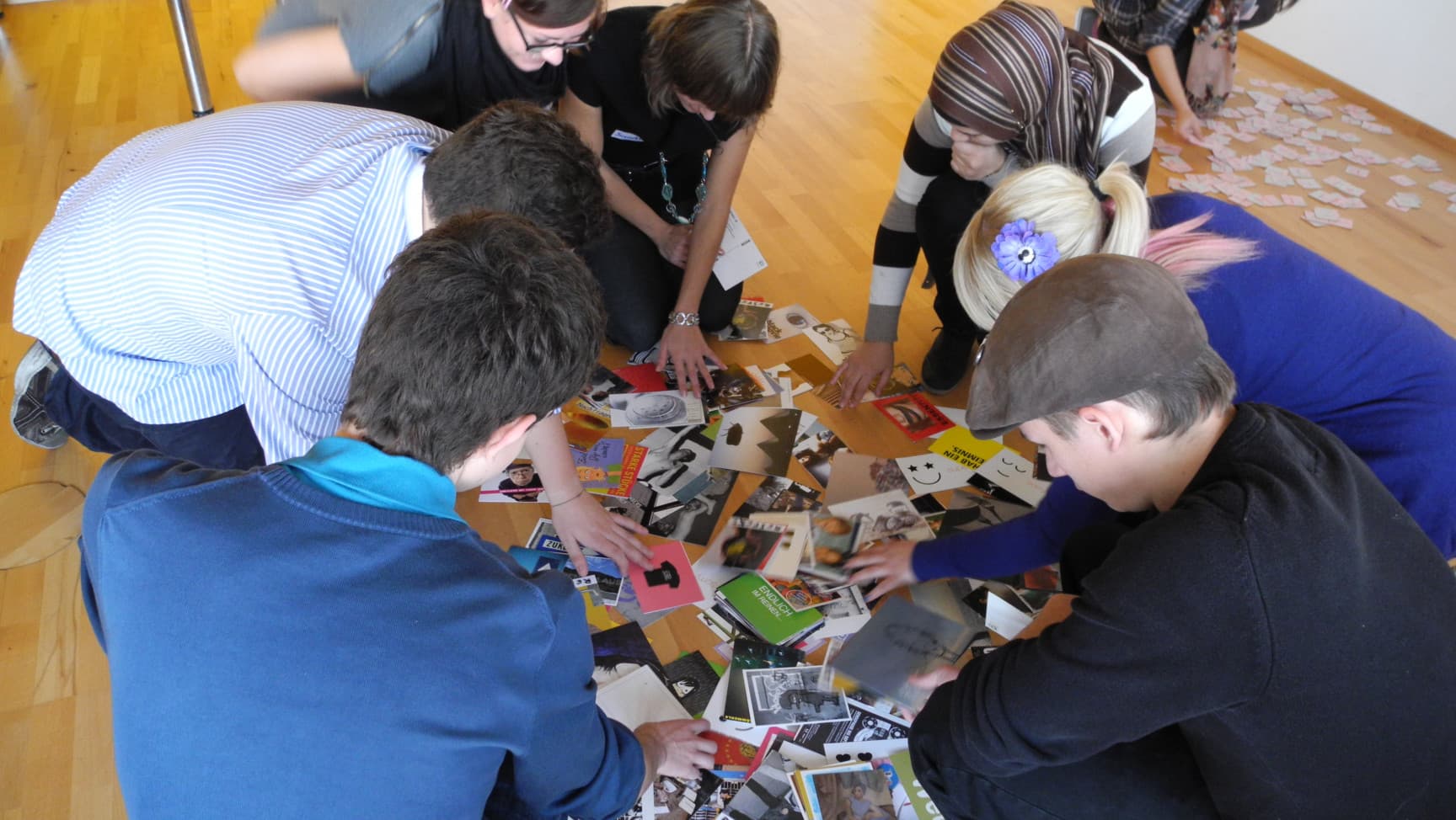 Jugendliche knieen am Boden und greifen nach vor ihnen verstreuten Postkarten.