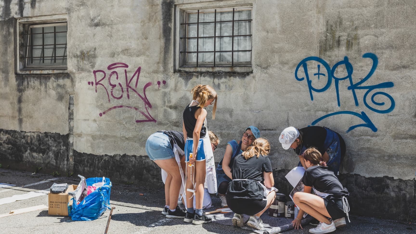 Eine Personenruppe hockt und steht vor einer Wand, vor ihnen auf dem Boden Farbkübel.