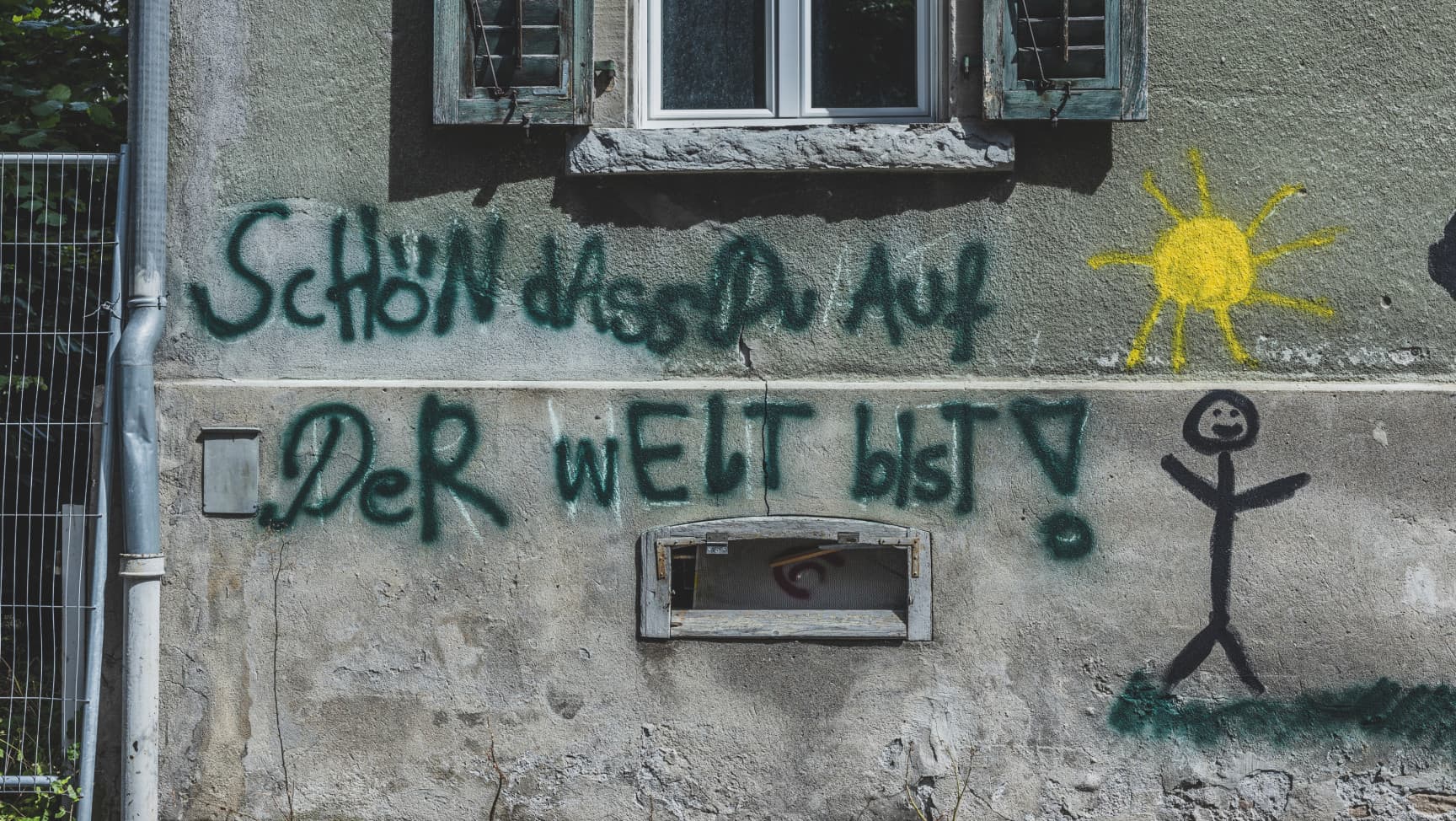 Auf einer Hauswand steht "Schön dass du auf der Welt bist!", daneben ist eine Sonne und eine Strichperson gemalt.