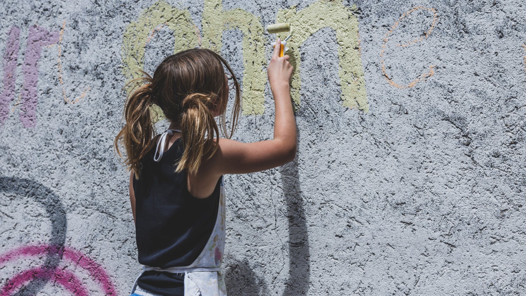 Eine jugendliche Person ist von hinten zu sehen, wie sie auf eine Wand mit einer Farbrolle gelbe Farbe aufträgt.