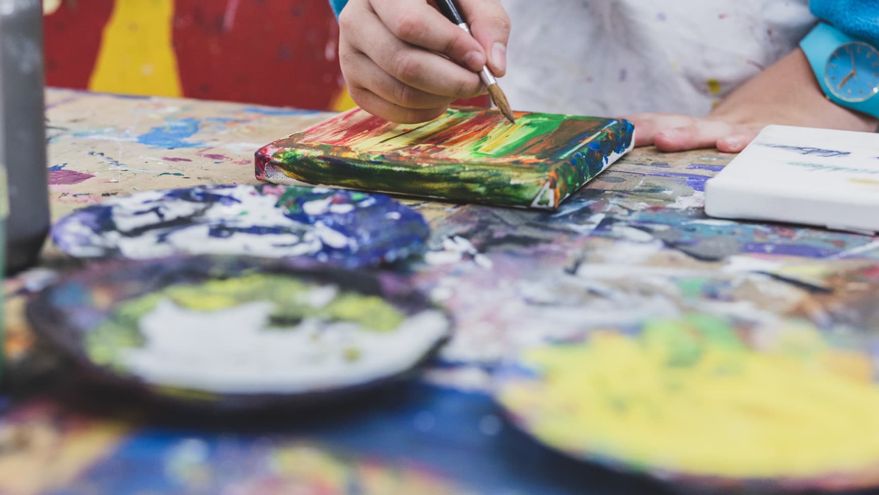 Hand einer jungen Person malt mit Pinsel ein Bild auf einem Werktisch.