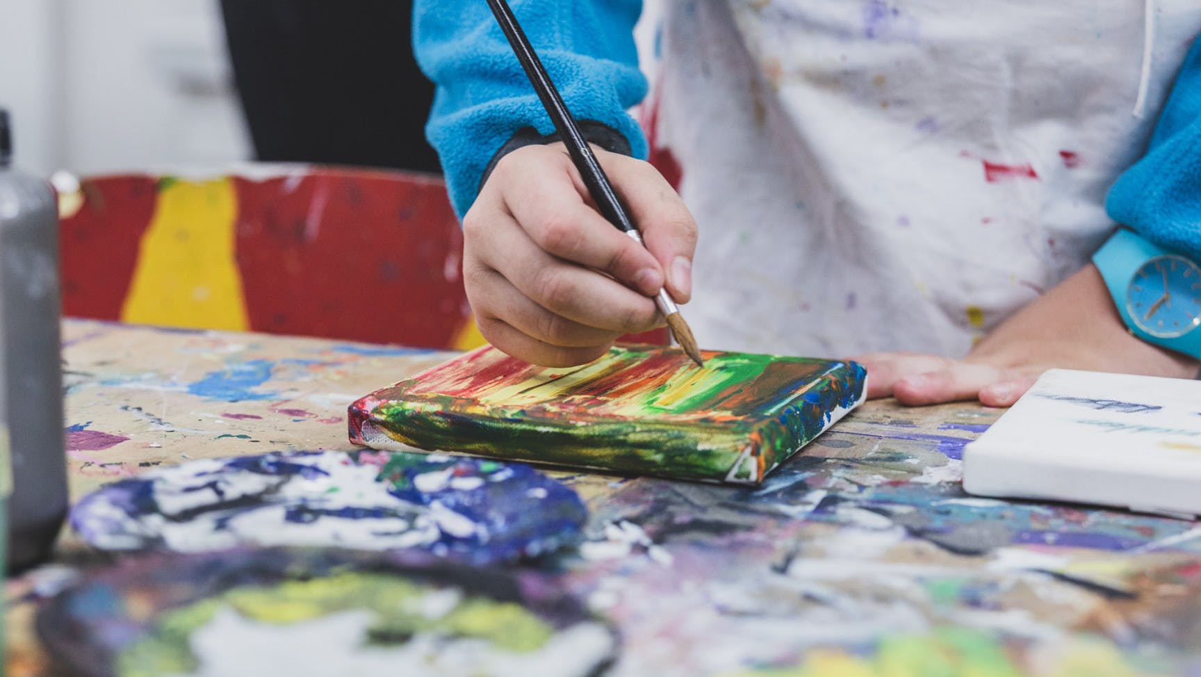 Hand einer jungen Person malt mit Pinsel ein Bild auf einem Werktisch.