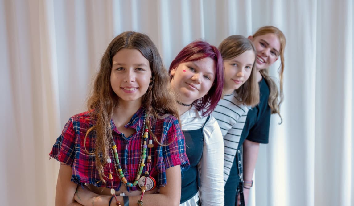 Vier FLINTA* Jugendliche stehen hintereinander und sehen lächelnd über die Schulter der vorderen Person nach vorne in die Kamera