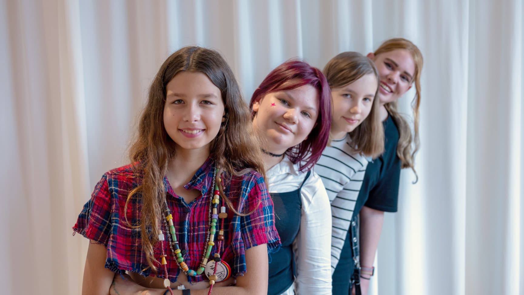 Vier FLINTA* Jugendliche stehen hintereinander und sehen lächelnd über die Schulter der vorderen Person nach vorne in die Kamera