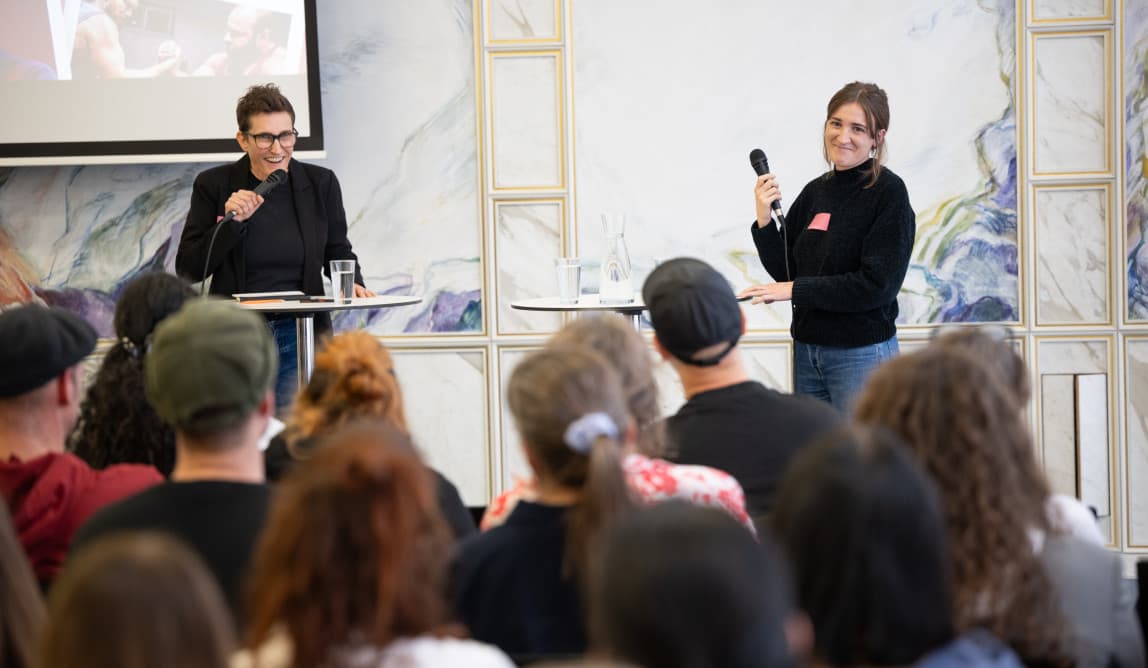 Zwei Sprecherinnen mit Mikrofonen stehen vor dem Publikum