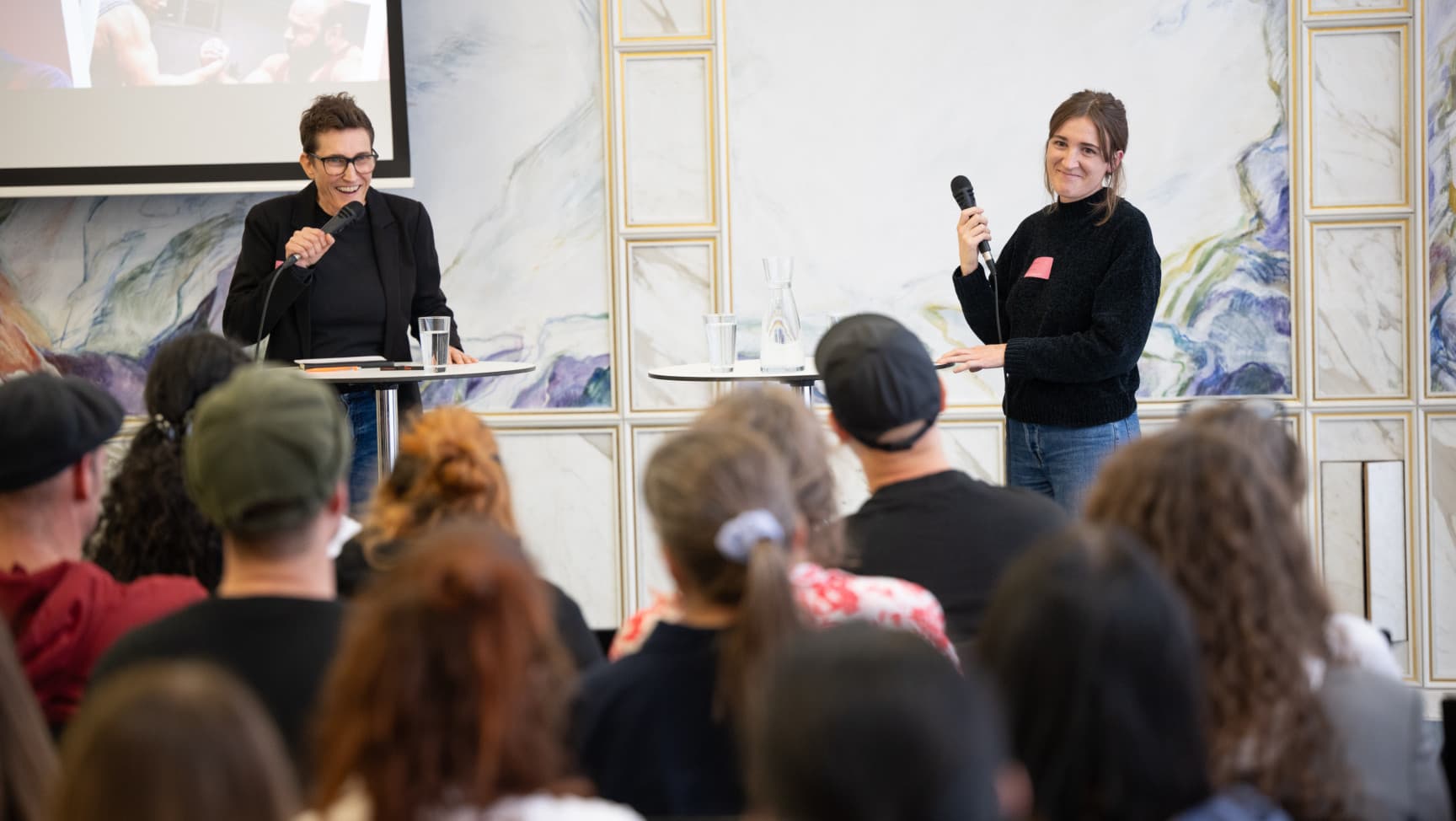 Zwei Sprecherinnen mit Mikrofonen stehen vor dem Publikum