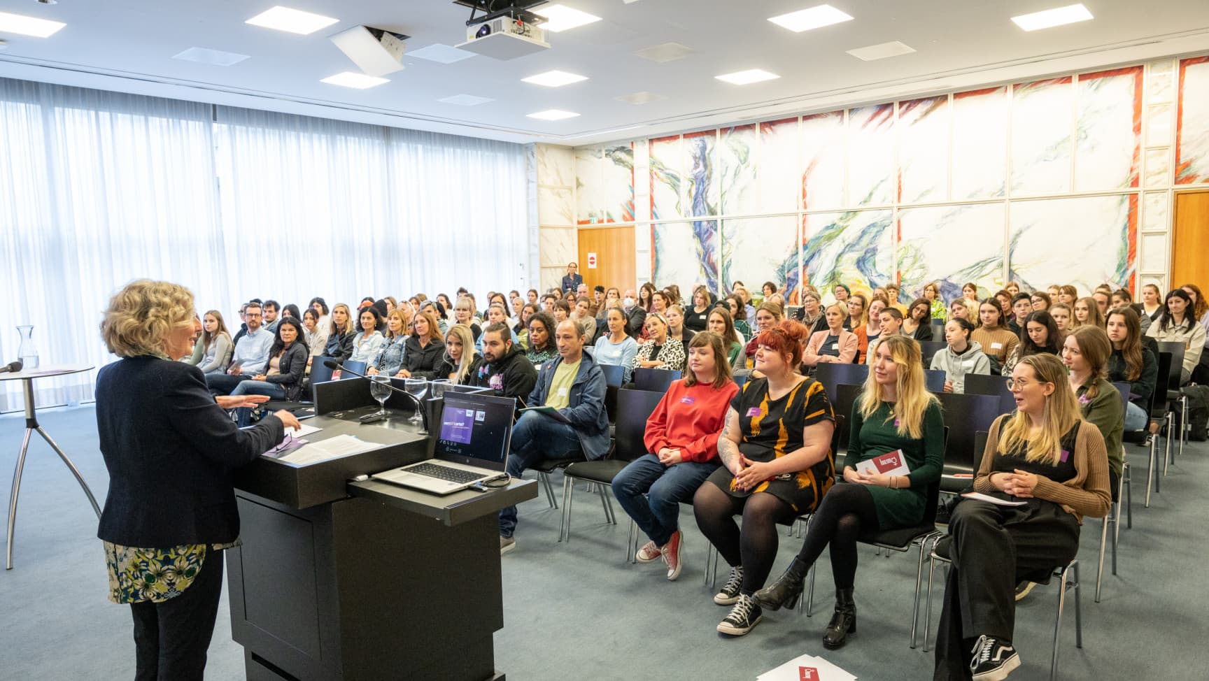 Sprecherin am Pult vor voll besetztem Saal mit Publikum.
