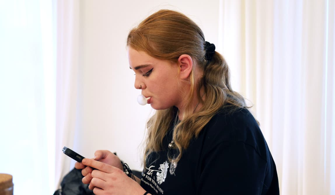 Eine jugendliche Person im Profil, die auf ihr Handy schaut und eine Kaugummiblase macht.