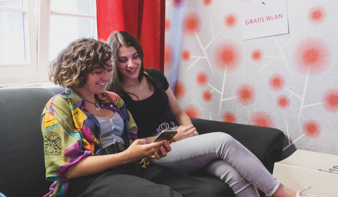 Zwei junge Frauen* sitzen auf einem Sofa und schauen lachend in ein Smartphone.