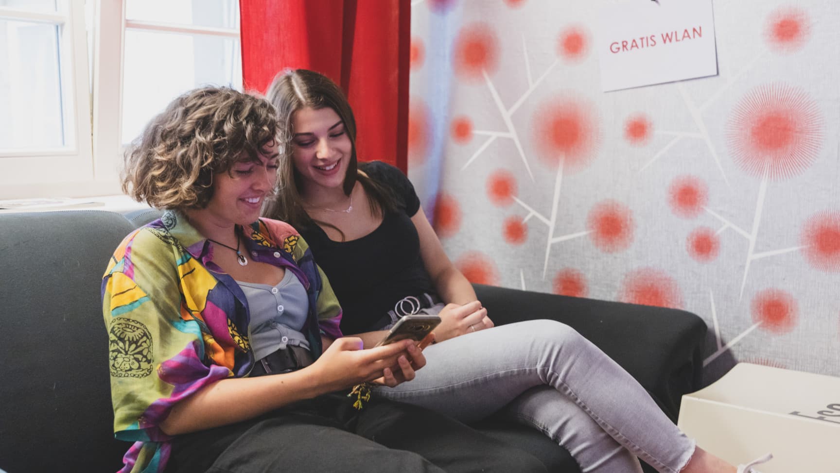 Zwei junge Frauen* sitzen auf einem Sofa und schauen lachend in ein Smartphone.