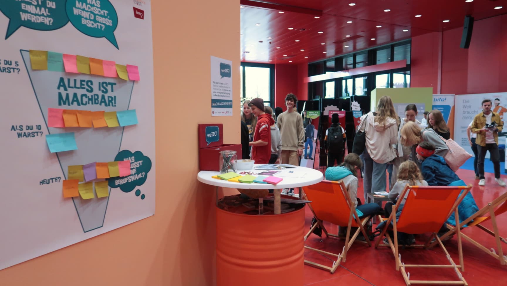 Wir sehen viele Jugendliche auf einer Messe, manche sitzen in Liegestühlen.