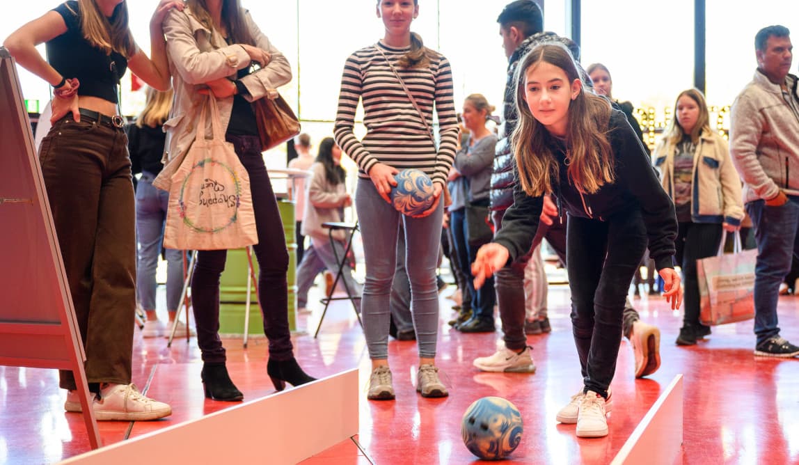 Ein Mädchen wirft eine Bowlingkugel in Richtung Kamera, dahinter stehen Menschen.