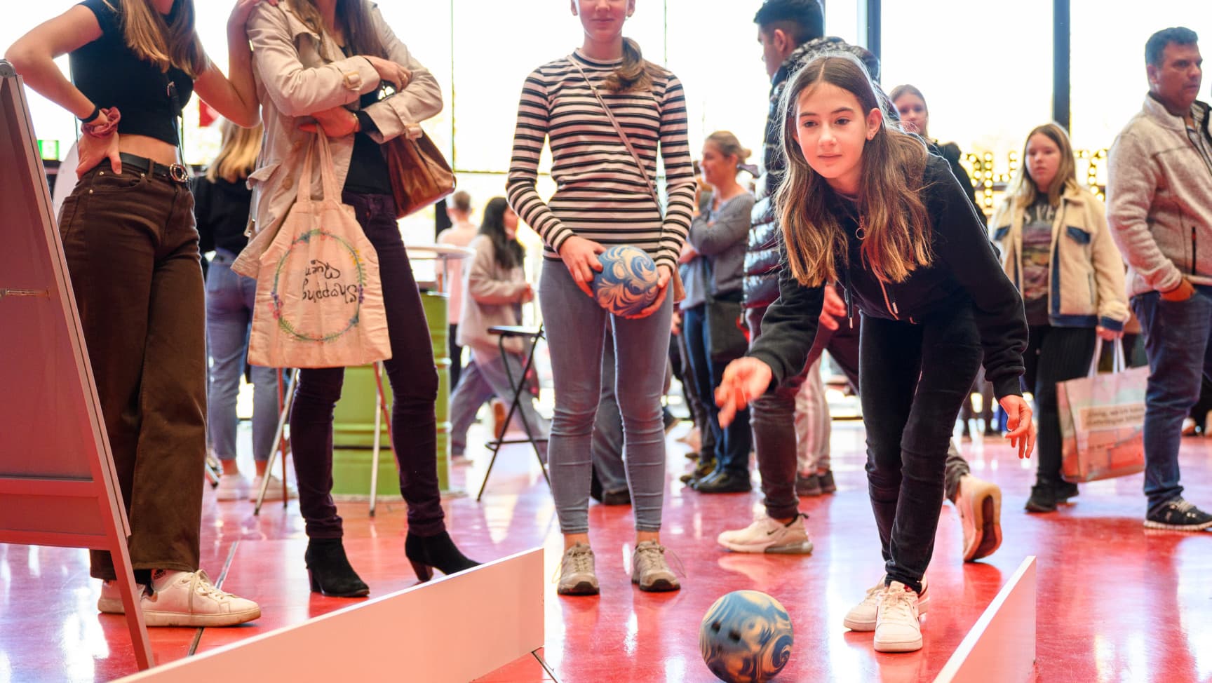 Ein Mädchen wirft eine Bowlingkugel in Richtung Kamera, dahinter stehen Menschen.