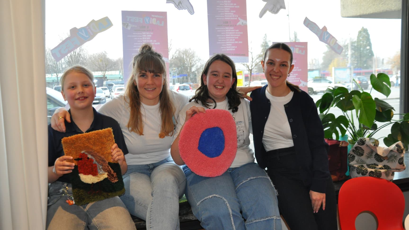 Vier junge Personen sitezn auf einem Fensterbank und schauen fröhlich, zwei von ihnen halten kleine Teppiche