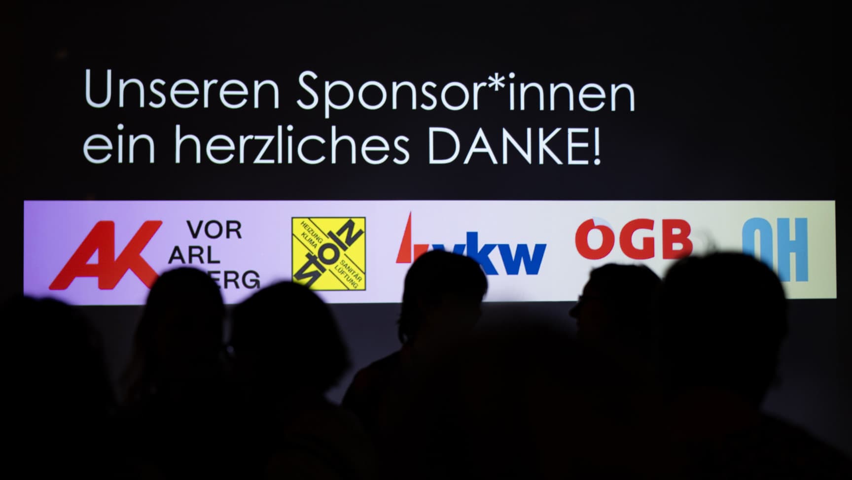 Menschen vor einer Wand mit Logos und dem Satz: Unseren Sponsor*innen ein herzliches Danke!