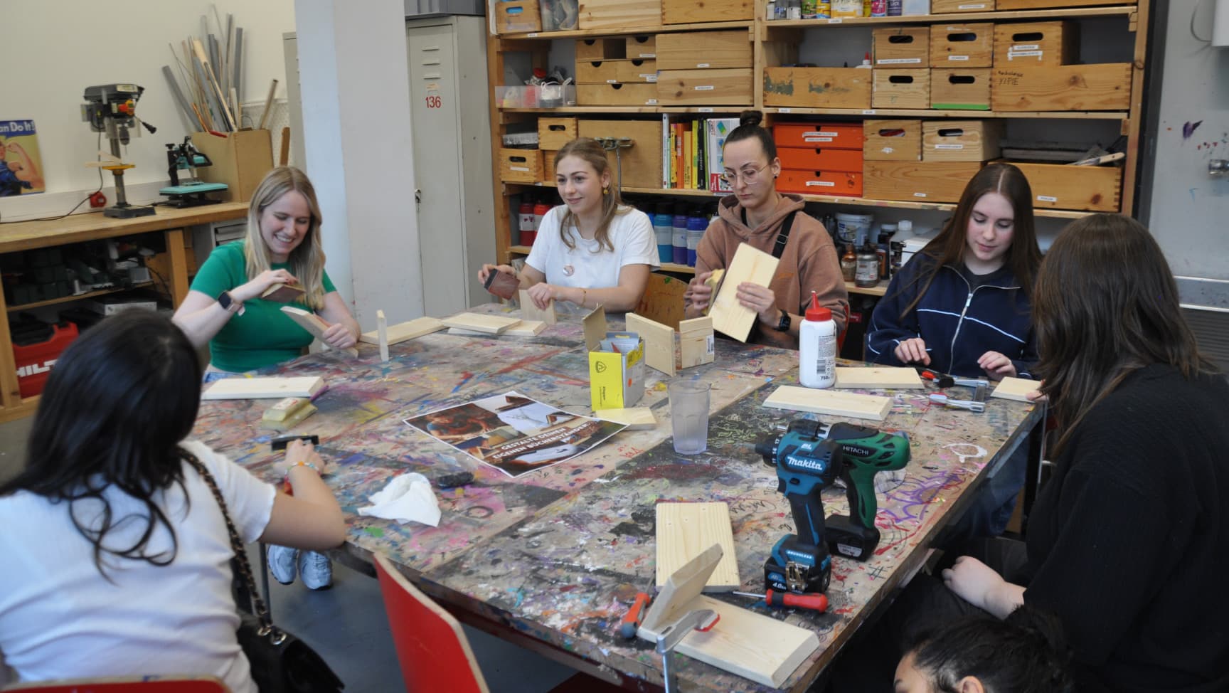 Mehrere Personen sitzen um einen Tisch in der Werkstatt und arbeiten mit Holz