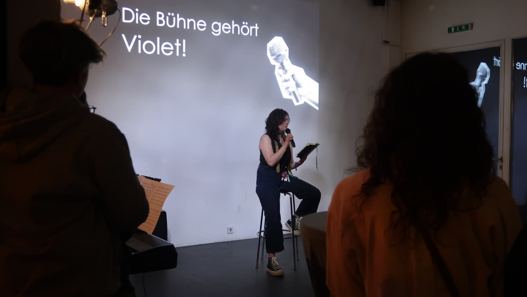 Person mit Mikrophon sitzt auf Hocker. Hinter ihr steht: Die Bühne gehört Violet!