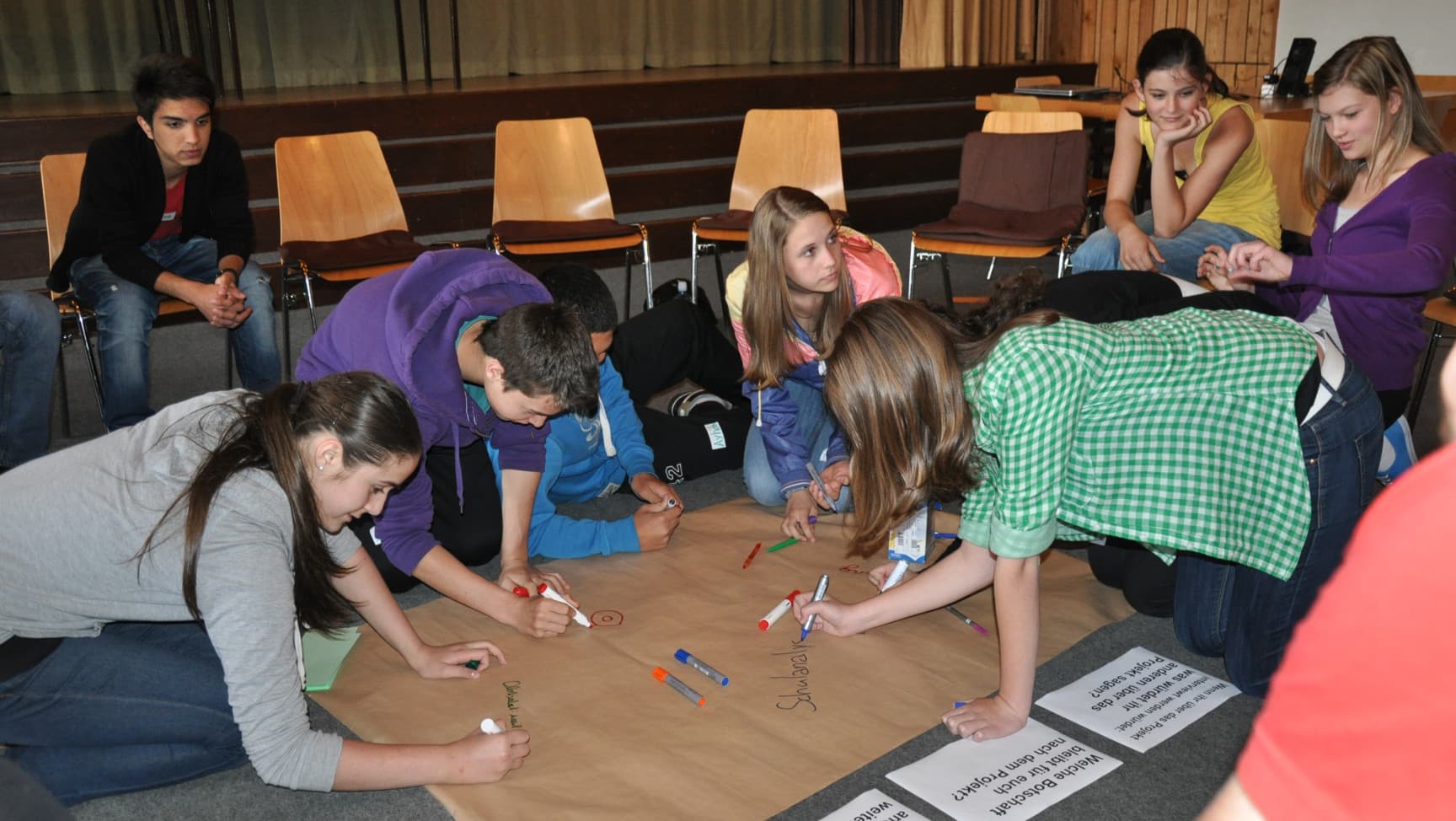 Workshopsituation mit Jugendlichen. Ein paar schreiben auf ein großes Blatt, das auf dem Boden liegt, ein paar sitzen auf Stühlen rundherum.
