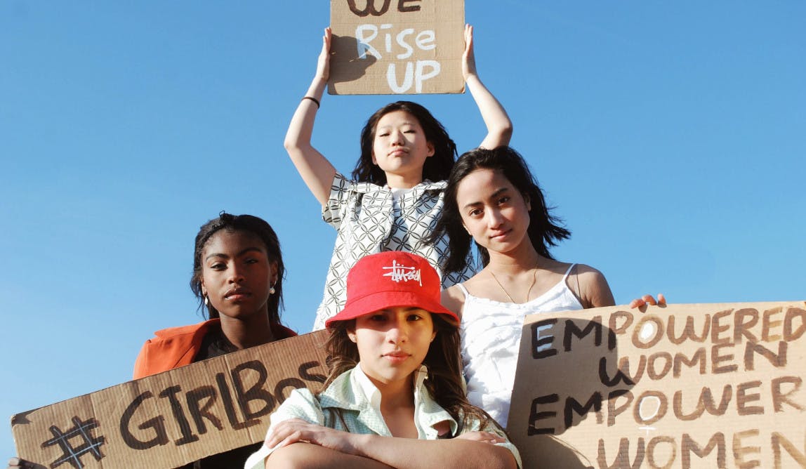 vier junge, weiblich gelesene Personen sind im Bild zusehen. Sie halten Kartons mit Sprüchen in die Höhe. Darauf steht: „We rise up“, #girlboss  „Empowered women empower women“
