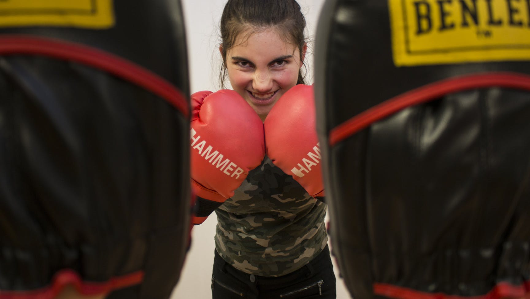 Zwischen zwei Kampfpratzen sieht man den Oberkörper einer weiblich gelesenen Person, die Boxhandschuhe trägt und sich diese in Kinnhöhe vors Gesicht hält.