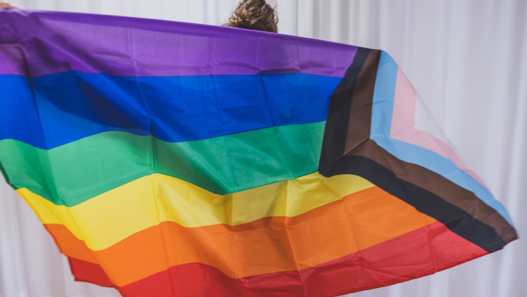 Eine Person hält eine große Progress Pride Flag mit beiden Armen weit ausgestreckt vor sich, das Gesicht ist nicht zu sehen.
