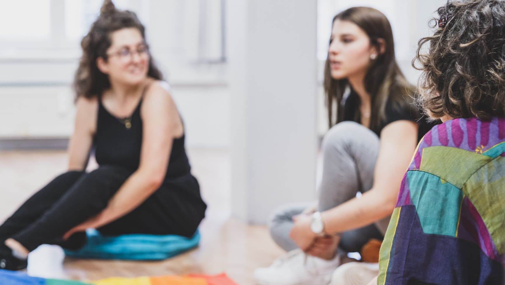 drei Personen sitzen auf dem Boden, in der Mitte liegt eine Regenbogenfahne
