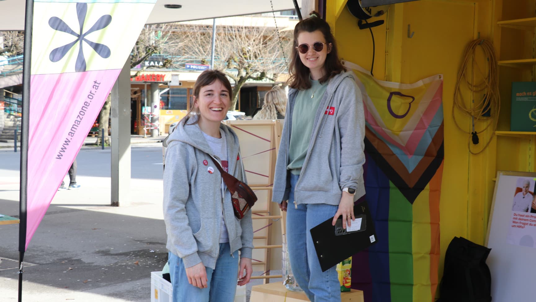 Zwei Frauen stehen auf einem Platz. Hinter ihnen eine Fahne und ein offener Holzraum mit Fahnen etc.