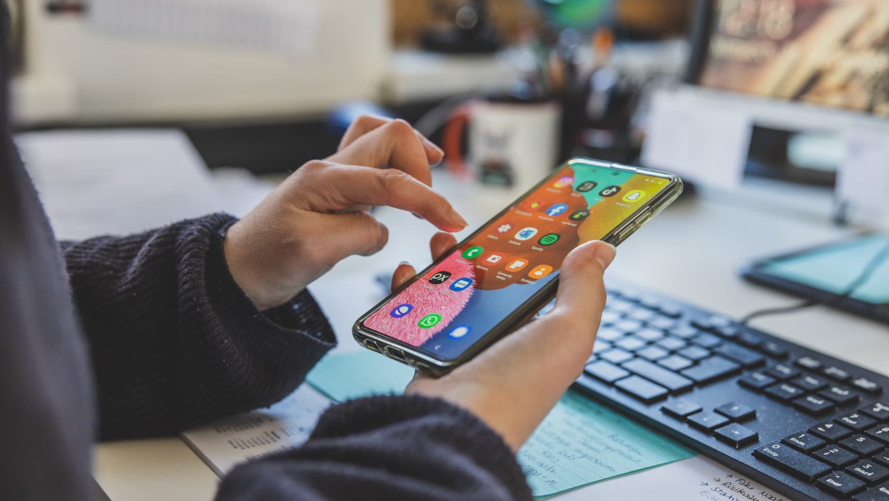 Nahaufnahme zwei Hände, die ein Smartsphone halten, im Hintergrund ist ein Schreibtisch mit Tastatur sichtbar