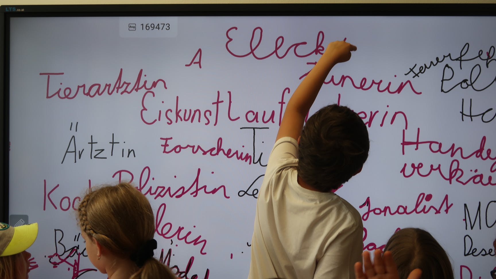 Ein Schüler* ist von hinten zu sehen, wie er etwas auf eine digitale Tafel schreibt.