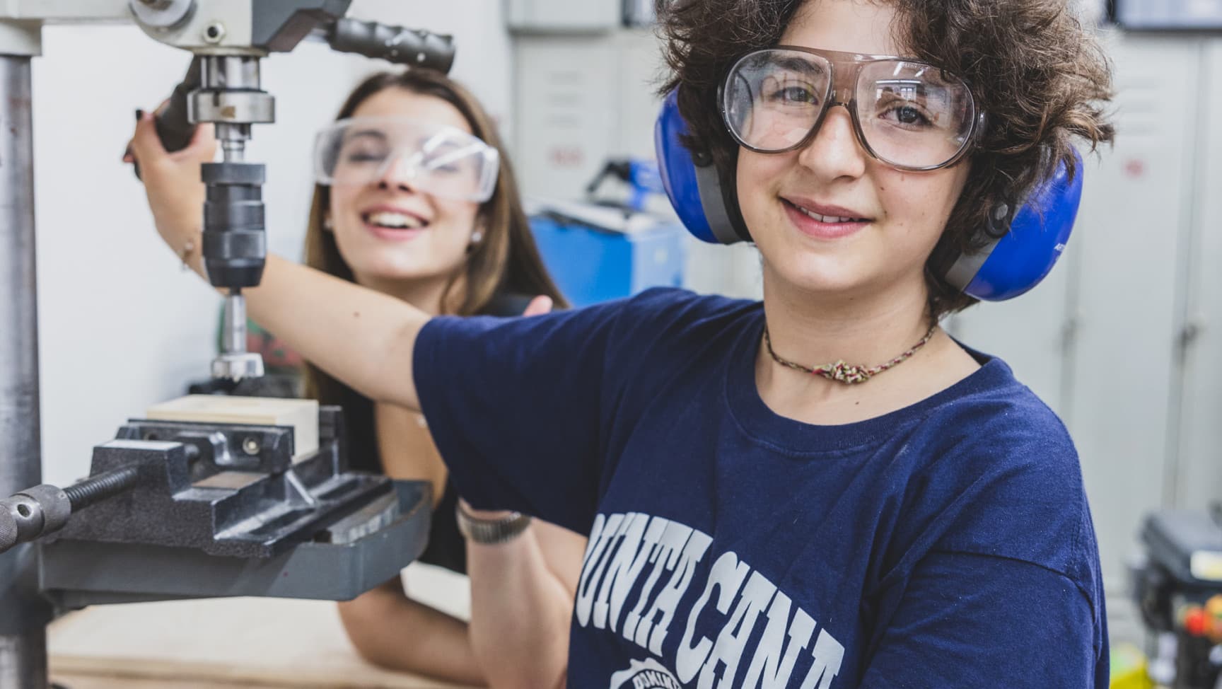 Zwei Jugendliche mit Schutzbrille und Lärmschutzhörern stehen vor einer Standbohrmaschine und lachen in die Kamera.